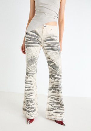 Kobieta w wysokich, rozszerzanych spodniach z abstrakcyjnym wzorem w odcieniach szarości i bieli, w połączeniu z czerwonymi butami na szpic oraz jasnoszarym topem.