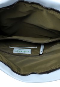 Bílá taška uvnitř s olivově zelenou podšívkou, obsahující zipovou kapsu a textilní štítek s názvem "SURFERRY GREEN LABEL." Hladká textura a zakřivený tvar.