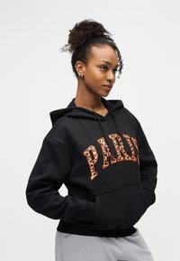 Νεαρή γυναίκα με σγουρά μαλλιά σε κότσο που φοράει μαύρο φούτερ με κουκούλα με τη λέξη «PARIS» σε γράμματα λεοπάρ, τα χέρια στις τσέπες, κοιτάζει δεξιά.