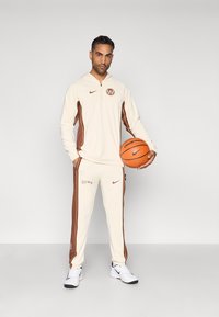 Kremo spalvos trenerių kostiumas su rudais akcentais, turintis užtraukiamąsias gobtuvą ir siaurėjančias kelnes. Komplekte yra krepšinis ir Nike ženklas ant aprangos.