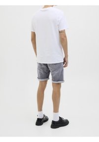 Mies, który ma na sobie białą koszulkę, rolowane szare dżinsowe szorty, białe skarpetki i czarne buty, stoi na tle jednolitego białego tła.