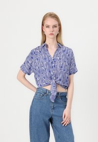 Blå kortermet skjorte med knyttet front og abstrakt hvitt bølgemønster, kombinert med høytlivs blå jeans.