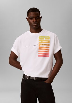 Mann trägt ein weißes Calvin Klein T-Shirt mit aufgerollten Ärmeln und vertikalem Logo im Farbverlauf von Gelb zu Rot, kombiniert mit schwarzer Hose und Gürtel.