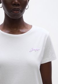 Fehér pamut póló kerek nyakkivágással, a bal mellrészen világoslila hímzett logóval. Rövid ujjú, sima textúrájú.