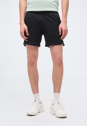 Muž v černých sportovních šortkách, bílých ponožkách a bílých teniskách stojící před jednoduchým světlým pozadím.