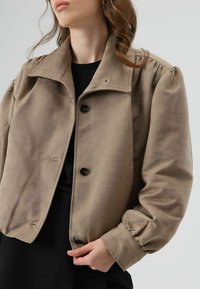 Beige cropped jacket with a collared neck, front button closure, and puffed sleeves. Made from soft fabric, featuring a smooth texture.