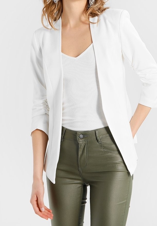 Femme portant un blazer blanc, un haut blanc sans manches et un pantalon ajusté vert olive avec boutons et poches visibles.