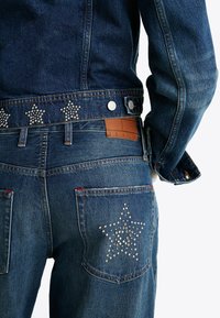 Džinsai su prigludusia forma, su žvaigždės formos studelėmis ant juosmens juostos ir galinės kišenės, taip pat su odiniu ženklu, pažymėtu "JEANS".