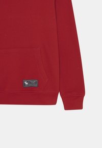Manica di una felpa rossa e parte inferiore del torso con tasca a marsupio e logo patch nero di Abercrombie & Fitch sull'orlo.