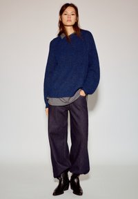 Oversized blauw gebreide trui met een ronde hals, gedragen over een grijs shirt, gecombineerd met donkerblauwe wijde spijkerbroek en zwarte enkellaarsjes.