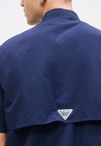 Άνδρας που φοράει μπλε σκούρο πουκάμισο PFG Performance Fishing Gear, εμφανιζόμενος από πίσω με το λαιμό και μέρος του προσώπου ορατά στο προφίλ.