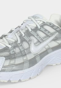 Λευκό και γκρι αθλητικό παπούτσι Nike με ύφασμα πλέγματος, δερμάτινες επικαλύψεις, λευκά κορδόνια και ορατό λογότυπο Nike Swoosh στο πλάι και στη γλώσσα.