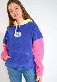 Pullover in pile con cappuccio giallo, con corpo blu e maniche rosa, una tasca frontale e un design di patch colorato sul petto.