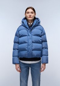 Giacca puffer blu con un design corto, caratterizzata da sezioni trapuntate orizzontali, un cappuccio e una chiusura con zip. Indossata sopra camicie sovrapposte.
