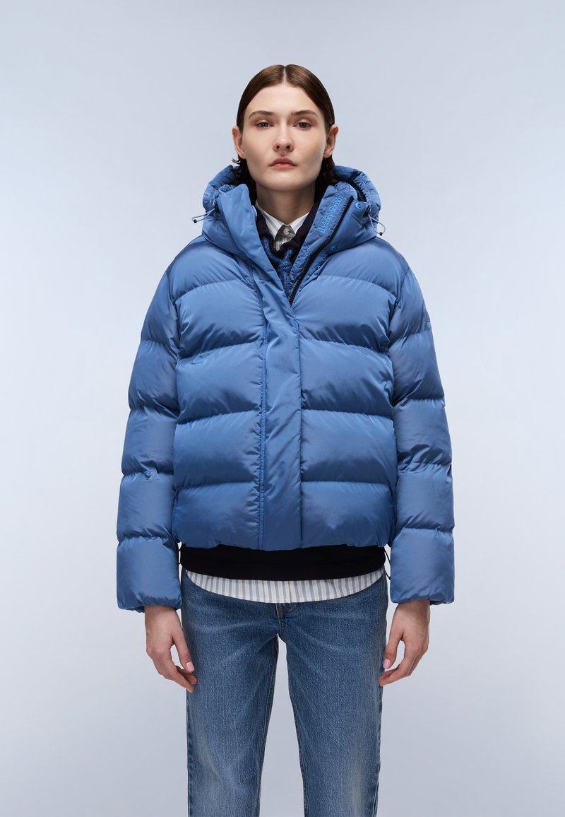 Giacca puffer blu con un design corto, caratterizzata da sezioni trapuntate orizzontali, un cappuccio e una chiusura con zip. Indossata sopra camicie sovrapposte.