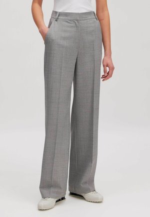 Frau trägt hoch taillierte, weit geschnittene graue Hose mit Bügelfalte vorne, weiße Sneakers und ein in die Hose gestecktes weißes Oberteil, steht mit einer Hand in der Tasche.