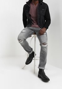Chaqueta de mezclilla negra con capucha, camiseta henley burdeos, jeans rasgados grises y botas negras hasta el tobillo. Sentado en un taburete blanco contra un fondo claro.