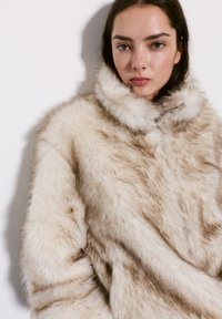 Faux fur jacket in beige and cream, featuring a high collar and a zip, with a soft, textured finish and uneven color patterns.