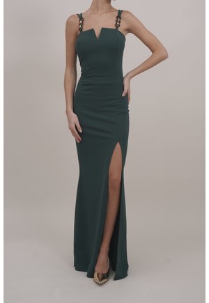 Kobieta w ciemnozielonej, bezrękawnikowej sukni z bocznym rozcięciem i ozdobnymi łańcuszkowymi ramiączkami, w połączeniu ze złotymi, spiczastymi szpilkami.