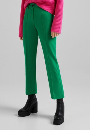 Pantalons droits verts avec des coutures avant, associés à un pull rose à côtes. Le look est complété par des bottes noires à talons épais.
