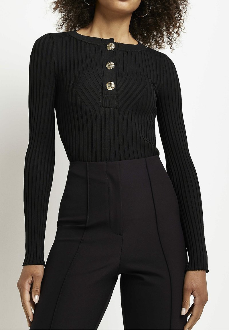 Femme portant un haut noir côtelé ajusté à manches longues avec trois boutons dorés et un pantalon noir taille haute, debout devant un fond blanc.