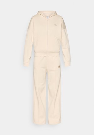 Beżowa bluza rozpinana na zamek i pasujące szerokie spodnie dresowe z subtelnym logo na klatce piersiowej i udzie, bez widocznych wzorów ani ozdób.