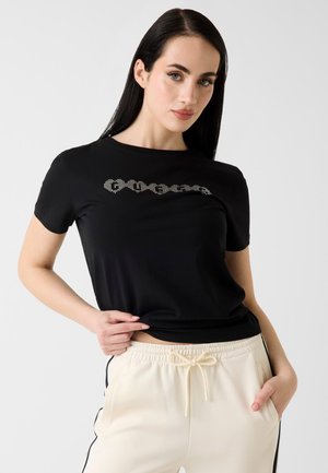 Femme aux longs cheveux foncés portant un t-shirt noir avec texte blanc et un pantalon crème à cordon avec des bandes noires sur les côtés.