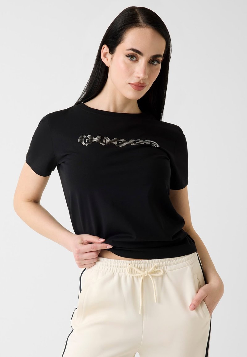 Femme aux longs cheveux foncés portant un t-shirt noir avec texte blanc et un pantalon crème à cordon avec des bandes noires sur les côtés.