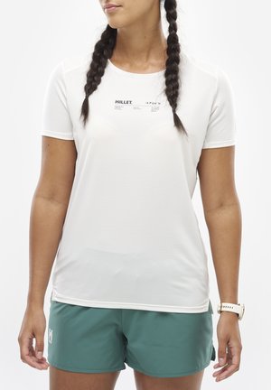 Femme aux cheveux tressés portant un t-shirt blanc Millet et un short de sport vert, avec une montre blanche au poignet gauche.
