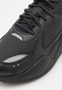 Černá sportovní obuv s koženým a síťovaným svrškem, texturovanými prvky, perforovanými detaily a plochými tkaničkami. Robustní gumová podrážka pro lepší trakci.