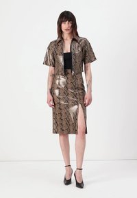 Veste courte à imprimé serpent avec une fermeture éclair avant, associée à une jupe midi assortie présentant une fente latérale, le tout en matériau brillant.