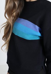 Suéter negro de punto con una franja de degradado horizontal que va del azul claro al azul oscuro en el pecho. Cuello y dobladillo acanalados.