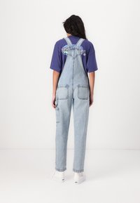 Salopette en denim bleu clair avec des bretelles ajustables, de grandes poches avant et une coupe décontractée, associée à un t-shirt violet et des chaussures blanches.
