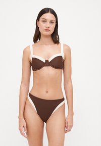 Brūns bikini komplekts ar krēmkrāsas akcentiem; krūšturis ar stieptiem stariem un platām siksnām; augsto vidukli un gludu tekstūru, minimālistisks dizains.