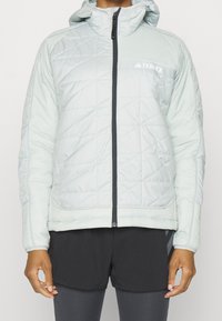 Veste matelassée bleu clair avec capuche et fermeture éclair noire. Présente un logo sur la poitrine et des poignets élastiques. Portée avec un short noir.