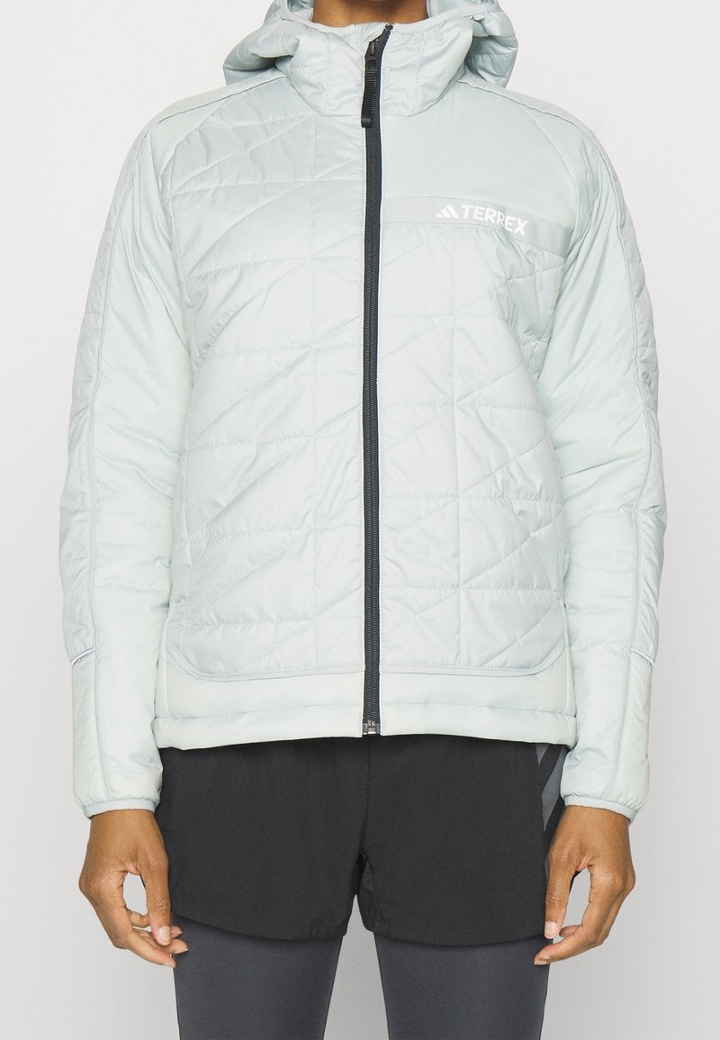 Veste matelassée bleu clair avec capuche et fermeture éclair noire. Présente un logo sur la poitrine et des poignets élastiques. Portée avec un short noir.