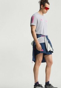 Muški sportaš u sivom majici s ružičastim trakama, tamnoplavim kratkim hlačama, vezanom jaknom, crvenim sportskim naočalama i crnim tenisicama za trčanje stoji bočno.