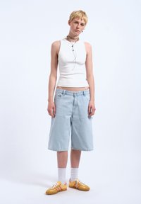 Top senza maniche bianco, pantaloni shorts di jeans blu chiaro che arrivano alle ginocchia, sneakers gialle con dettagli rosa e calzini bianchi alla caviglia.