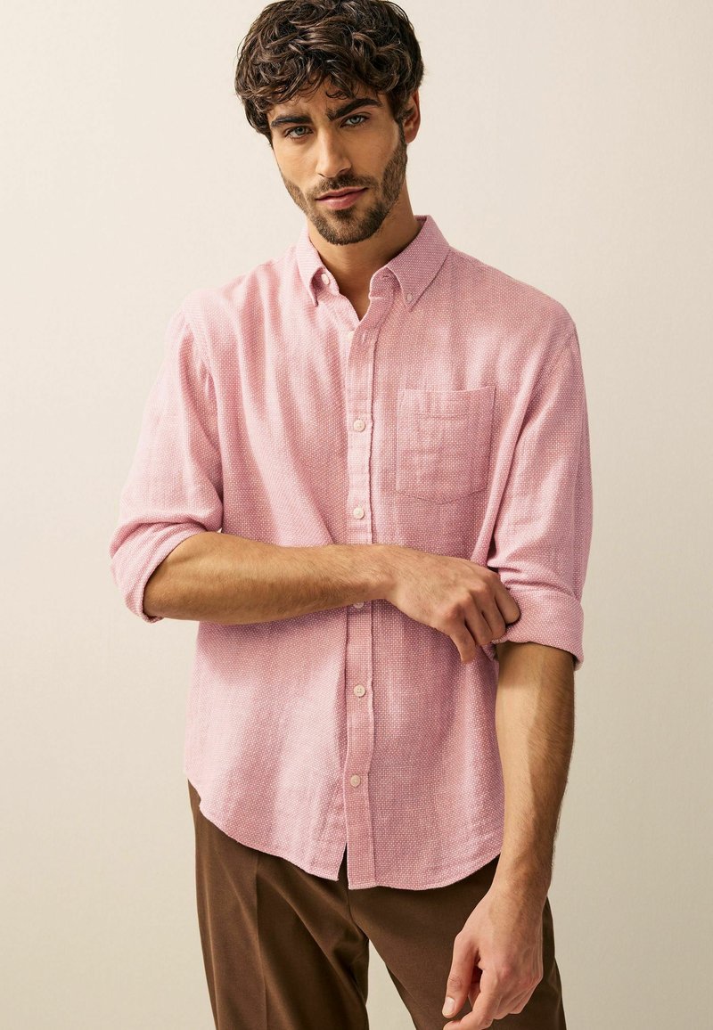 Camisa de lino rosa claro con un patrón texturizado, cuello abotonado y un bolsillo en el pecho. Mangas enrolladas y dobladillo curvado. Pantalones marrones.