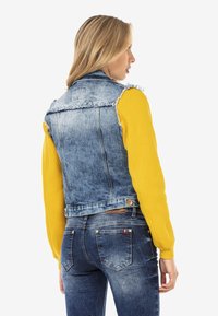 Veste en denim avec des bords effilochés, lavage bleu clair, associée à une chemise à manches longues jaune. Dispose d'une taille ceinturée et de poches classiques.