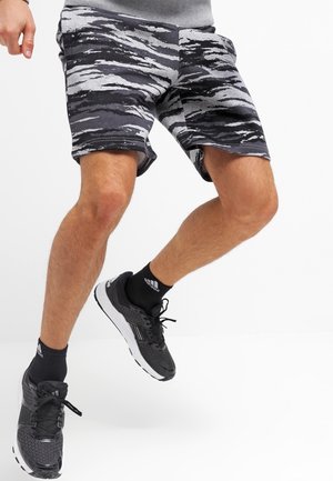 Personne portant un short noir et gris à motifs, des chaussettes noires Adidas et des chaussures de sport noires, saisie en plein saut sur fond blanc.