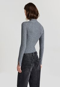 Frau mit braunen Haaren, die ein enges, graues Rippenshirt mit langen Ärmeln und hochtaillierte, ausgebleichte schwarze Jeans trägt und vor einem schlichten hellen Hintergrund steht.