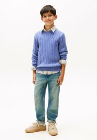 Maglione a collo alto in lana leggera color viola, camicia a scacchi bianca e grigia sotto, jeans di denim blu e sneakers beige con dettagli testurizzati.