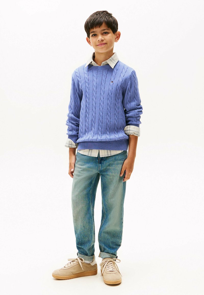 Maglione a collo alto in lana leggera color viola, camicia a scacchi bianca e grigia sotto, jeans di denim blu e sneakers beige con dettagli testurizzati.