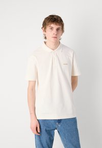 Krems krāsas polo krekls ar apkakli un divām pogām, ar smalku Calvin Klein logotipu brūnā krāsā. Valkāts ar ziliem denima džinsiem. Mīksta auduma.