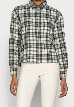 Personne portant une chemise à carreaux boutonnée à manches longues blanche, noire et marron avec un pantalon de couleur claire, debout devant un fond uni.