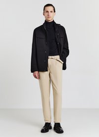 Giacca nera con zip e due tasche sul petto, indossata sopra un dolcevita nero. Pantaloni beige e scarpe nere completano l'outfit.