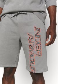Shorts en coton gris avec une taille élastique et un cordon de serrage. Présente un grand texte orange et un motif camouflage sur le côté gauche.