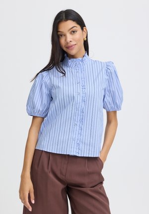 Sieviete ar gariem tumšiem matiem, tērpusies gaiši zilā, īsroku krekliņā ar vertikālām svītrām un volānu detaļām, valkājot brūnas bikses, stāv pret vienkāršu fona sienu.