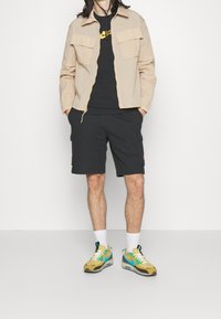 Giacca leggera beige con due tasche frontali, t-shirt nera, pantaloni corti scuri e sneakers giallo-blu con dettagli bianchi.